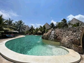 Una piscina cristalina con una pared de piedra y palmeras al fondo.