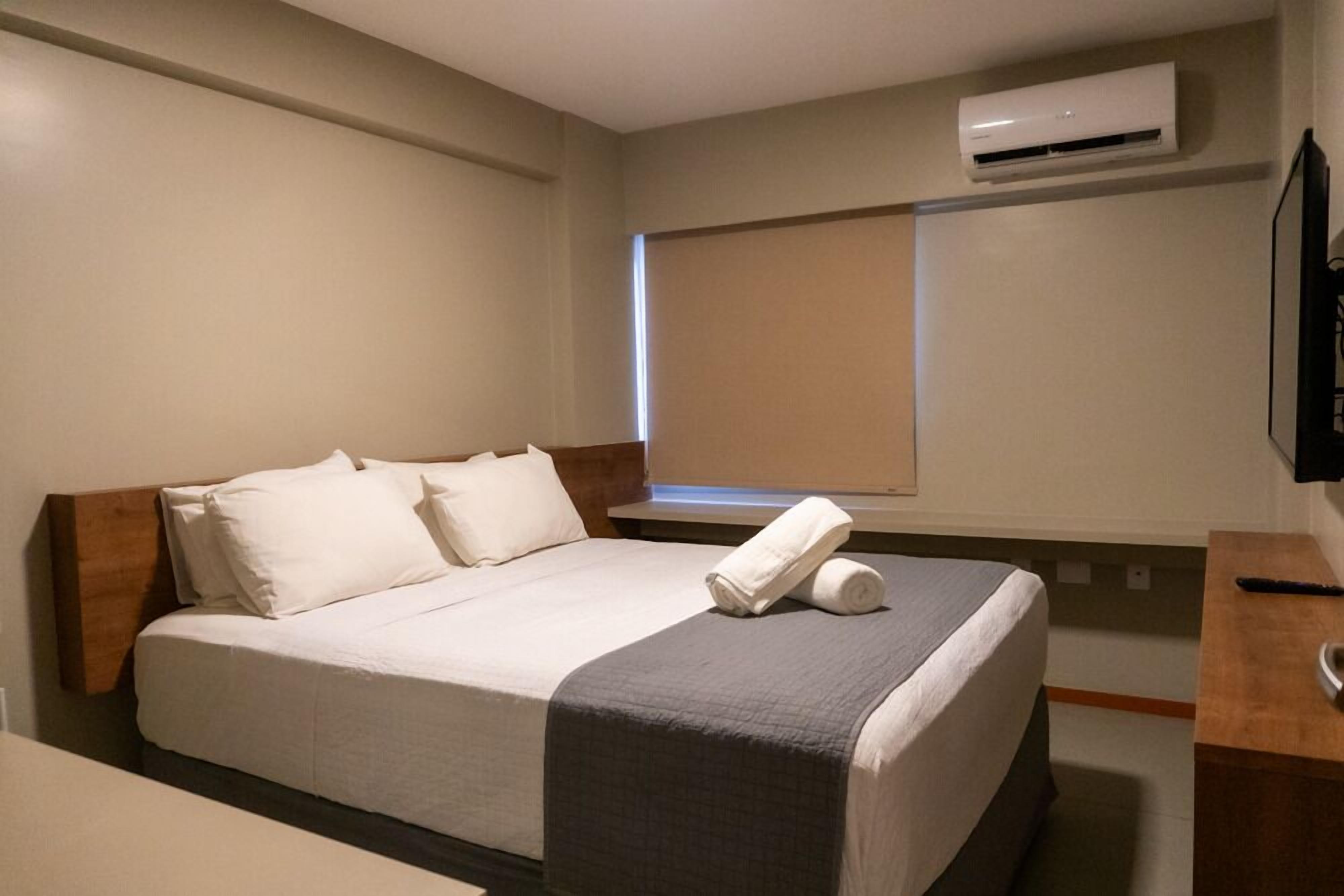 A hotel room with a bed, a wooden headboard, a bedside table, a TV, and an air conditioning unit.