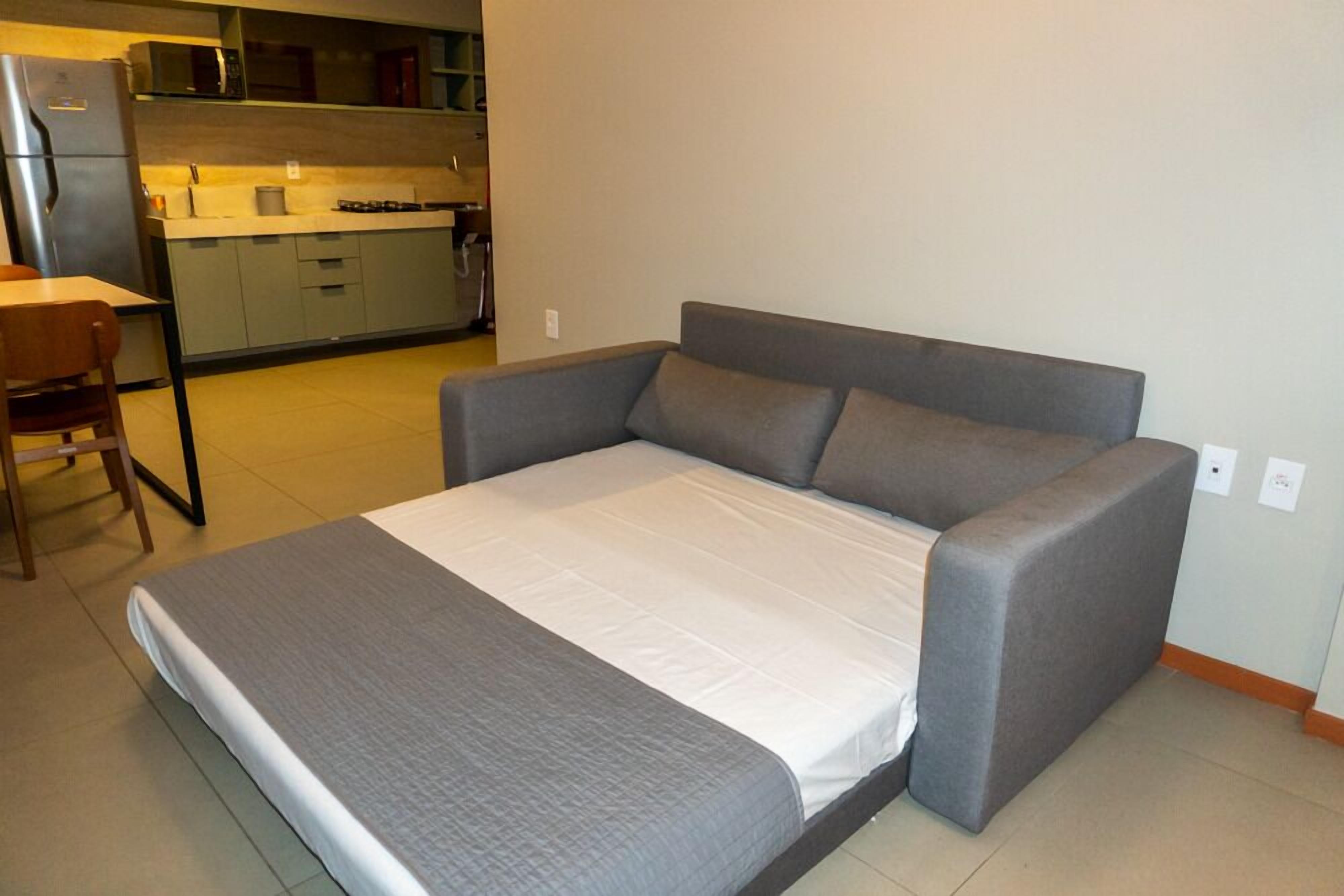 A compact bed with a grey mattress and pillows, a wooden chair, and a kitchenette in the background.