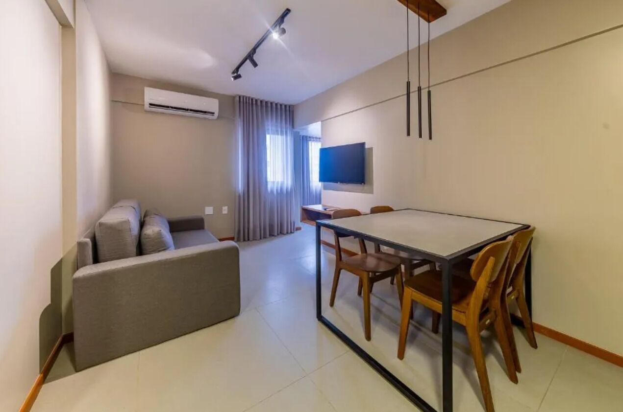 A modern living room with a sofa, a dining table with chairs, a flat-screen TV mounted on the wall, and an air conditioning unit.