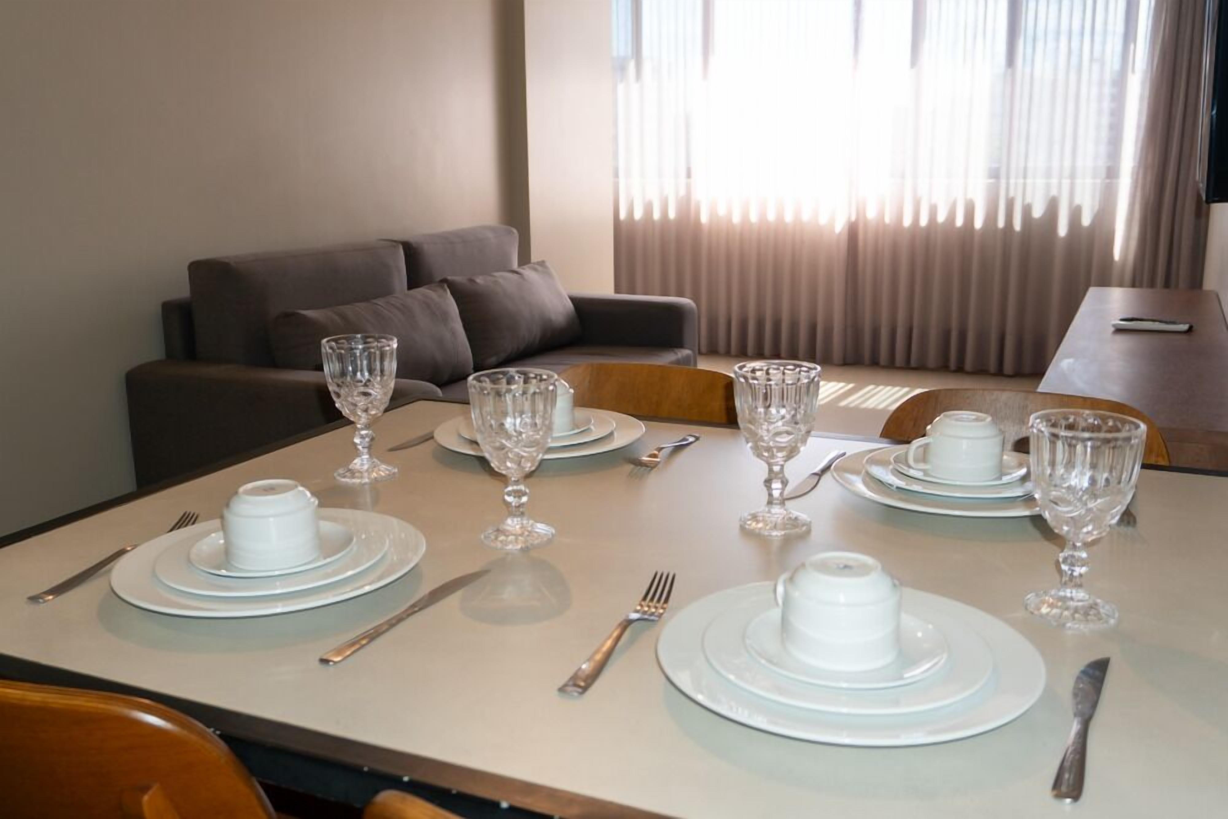 A dining table set with plates, glasses, and cutlery, with a sofa and curtains in the background.