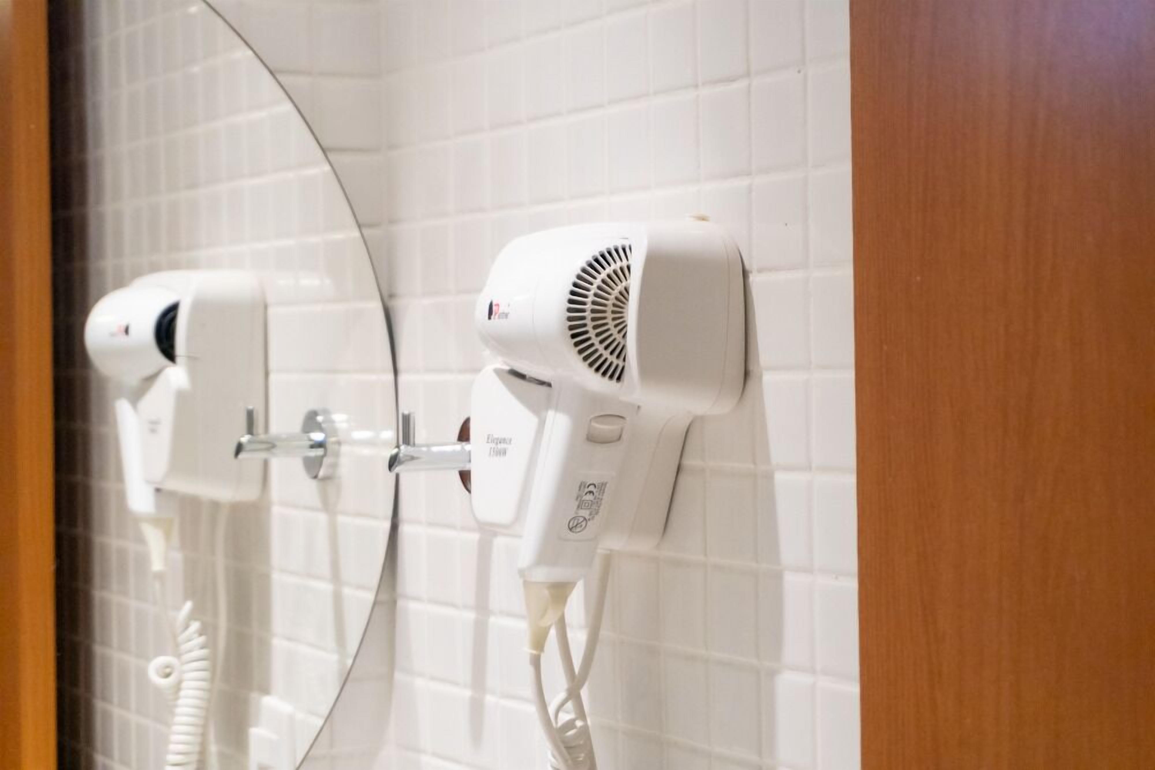 Un baño con paredes de azulejos blancos, una puerta de madera y dos secadores de pelo fijados a la pared.