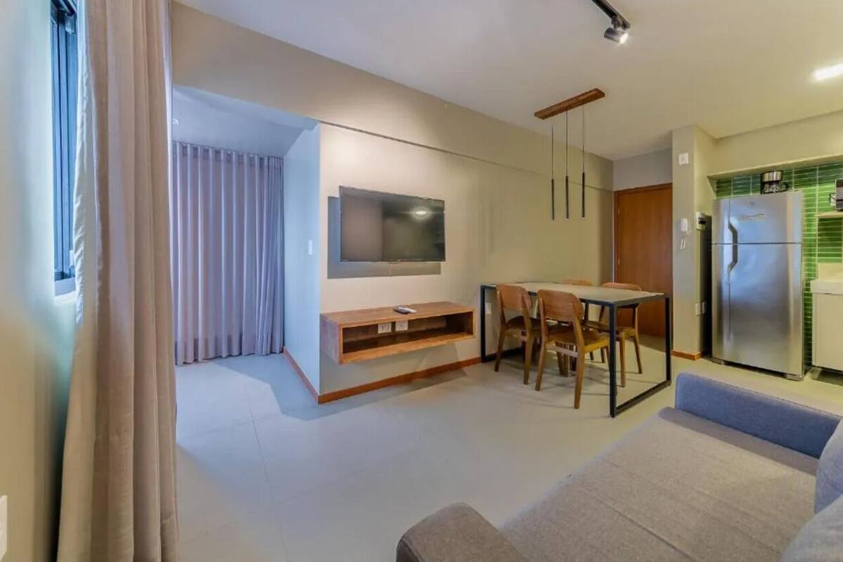 A modern living room with a flat-screen TV, a dining area with a table and chairs, and a kitchen with a refrigerator.