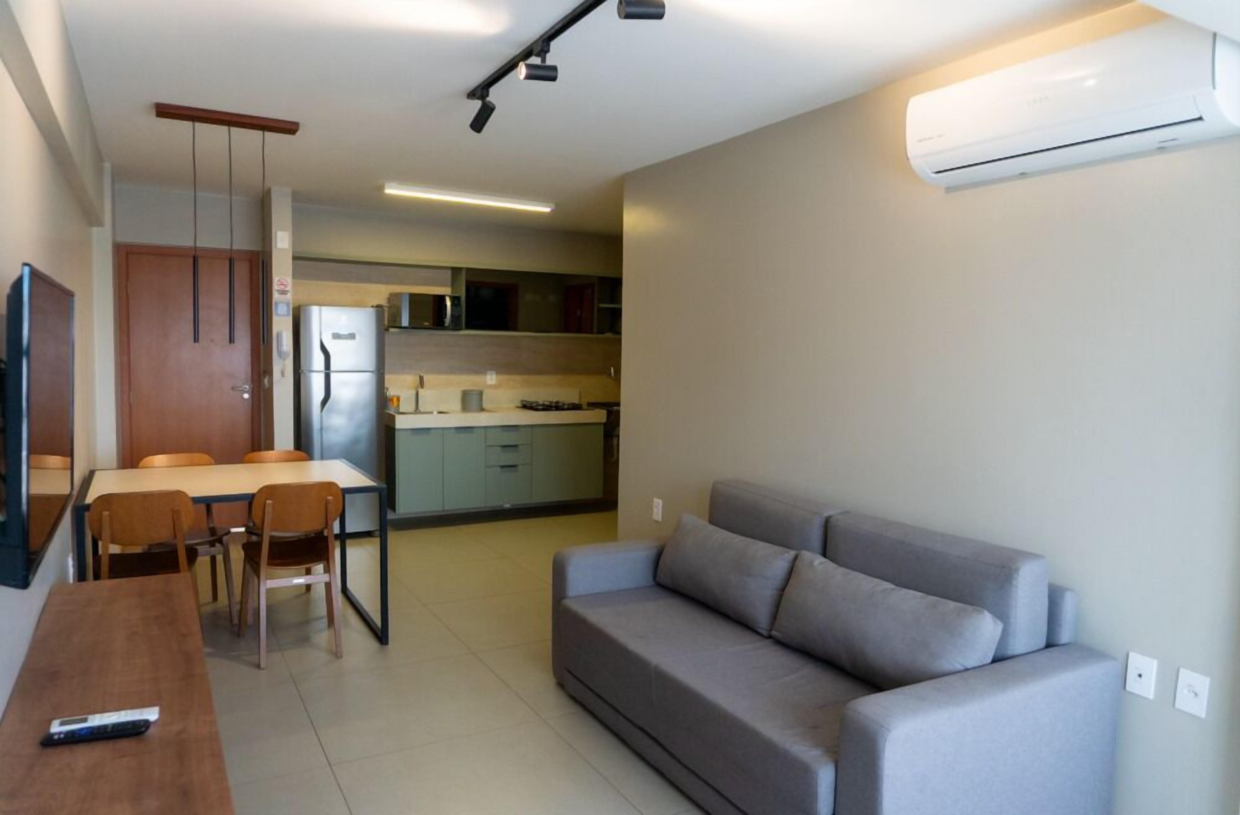 A modern living room with a sofa, a dining table with chairs, a kitchen area in the background, and an air conditioning unit mounted on the wall.
