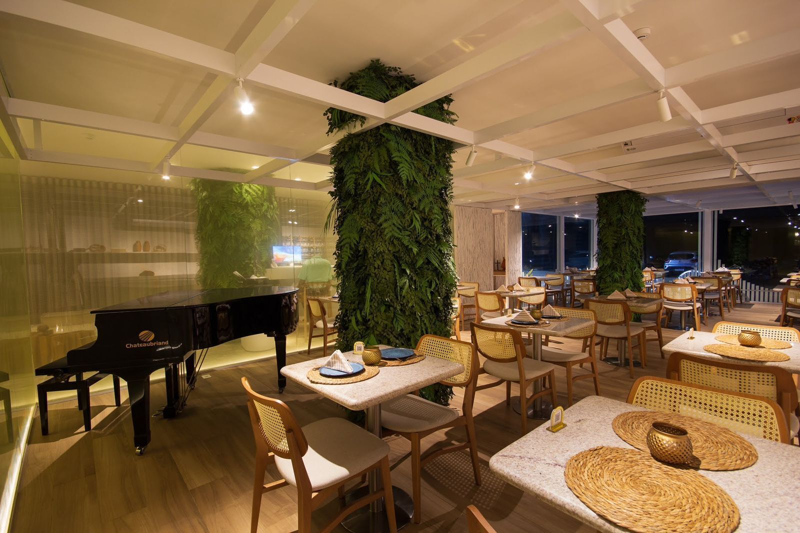 A restaurant with a piano, tables with woven placemats, and large green vertical gardens.