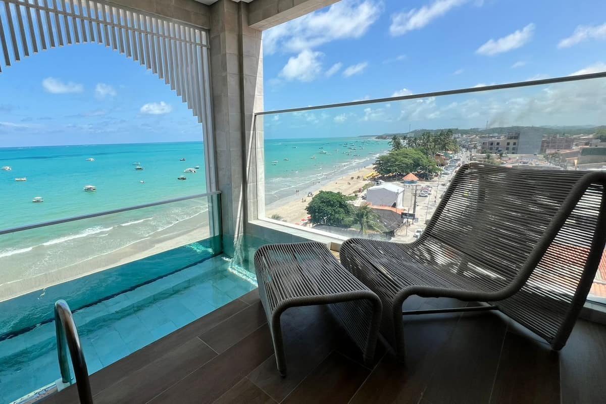 Un balcon offrant une vue sur une plage et l’océan, avec deux fauteuils en osier et une balustrade en verre.