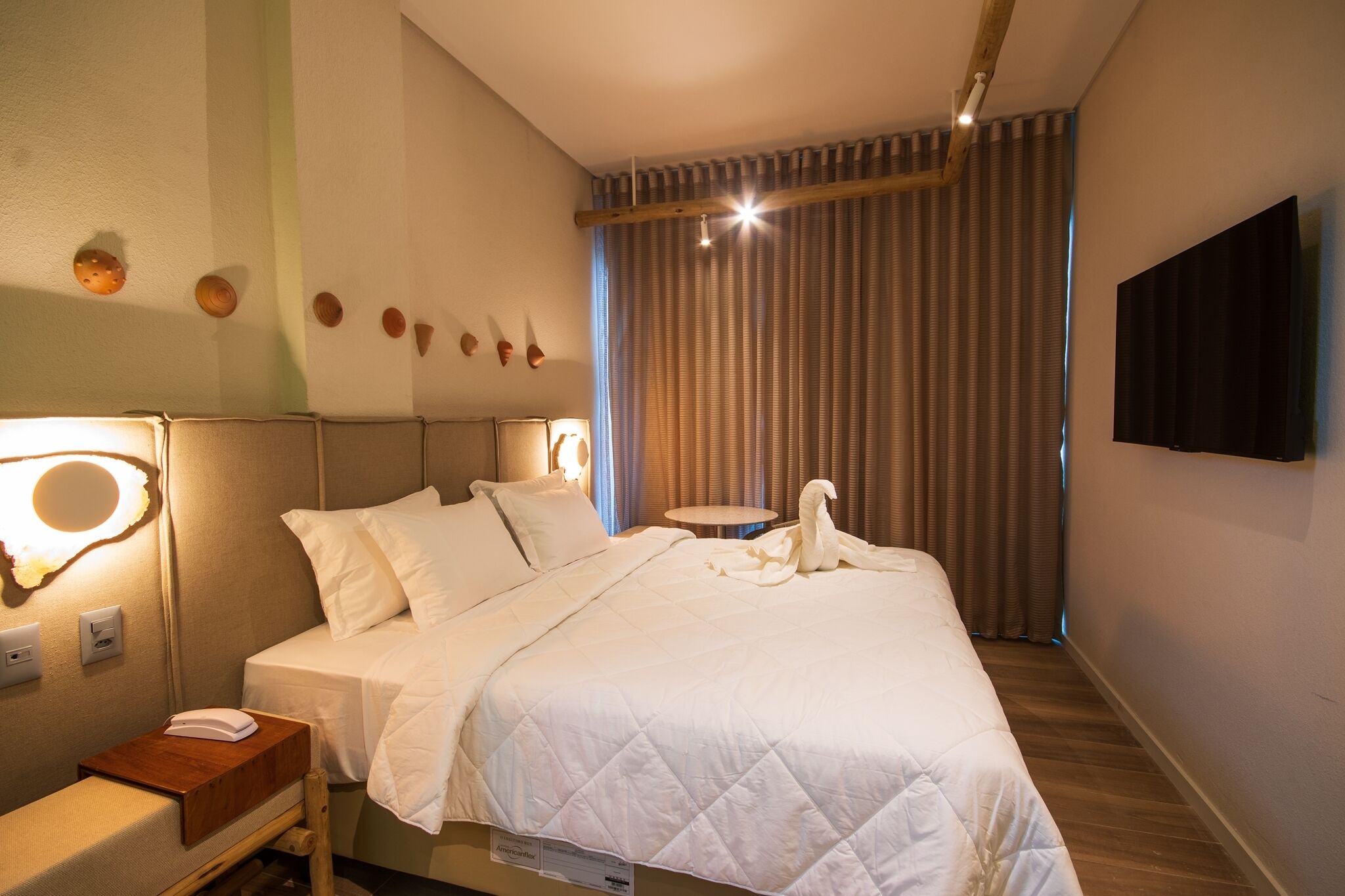 Un lit bien fait, avec une couette blanche matelassée, une table de chevet sur laquelle se trouvent un livre et une lampe, et un téléviseur fixé au mur.