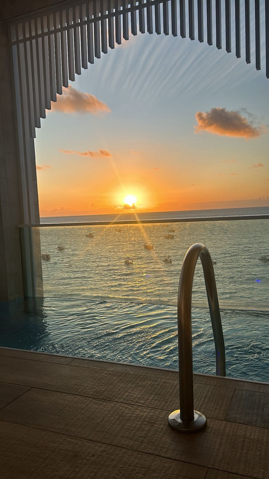 Vue du coucher de soleil sur l’océan, prise depuis un espace piscine intérieur doté d’une rambarde en métal.