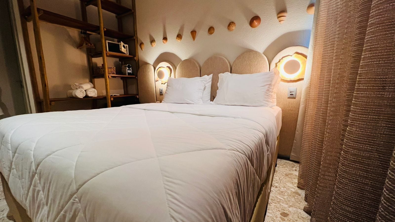 Una cama bien tendida con edredón blanco, cabecera de madera con luces circulares, y un estante con toallas dobladas.