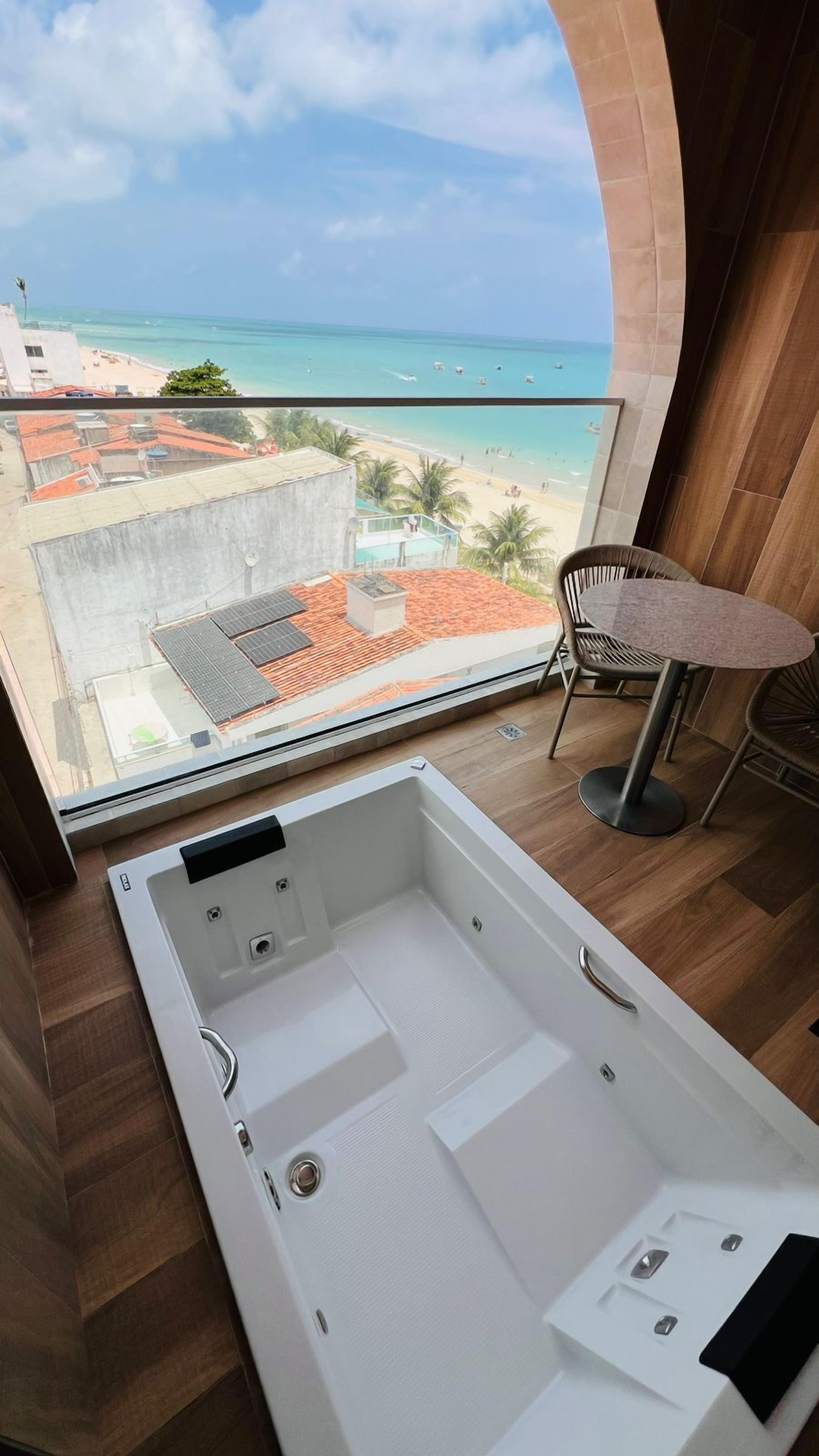 Un baño moderno con una bañera grande, vistas a una playa y una pequeña mesa con sillas.