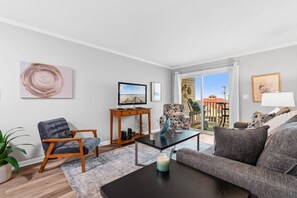 Een moderne woonkamer met een flatscreen televisie, een bank, een fauteuil en een salontafel. Er staat een plant, een lamp en er hangt kunst aan de muren.