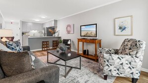 Una sala moderna con un sofá, una mesa de centro y un sillón estampado. Un televisor está sobre un mueble de madera, y se aprecia una cocina al fondo.