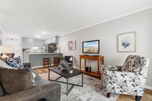 A modern living room with a sofa, coffee table, and patterned armchair. A television is on a wooden stand, and there is a kitchen in the background. - Beachside Hideout- Pool- 3 Bds- Elevator (North Myrtle Beach)