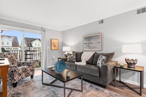 A living room with a sofa, coffee table, and side tables, a large window with a view of houses, and a painting on the wall. - Beachside Hideout- Pool- 3 Bds- Elevator (North Myrtle Beach)