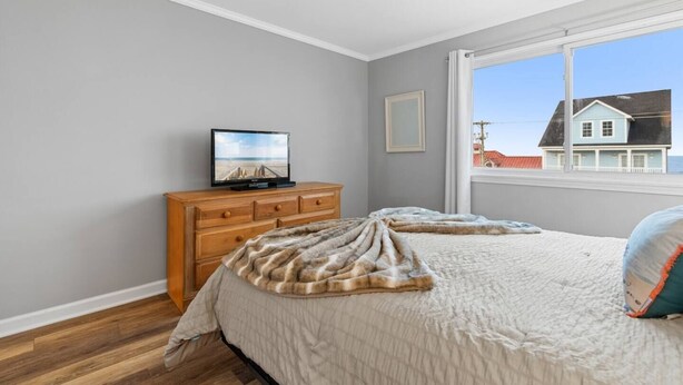 Un dormitorio con cama, cómoda, televisor y ventana con vista a una casa.