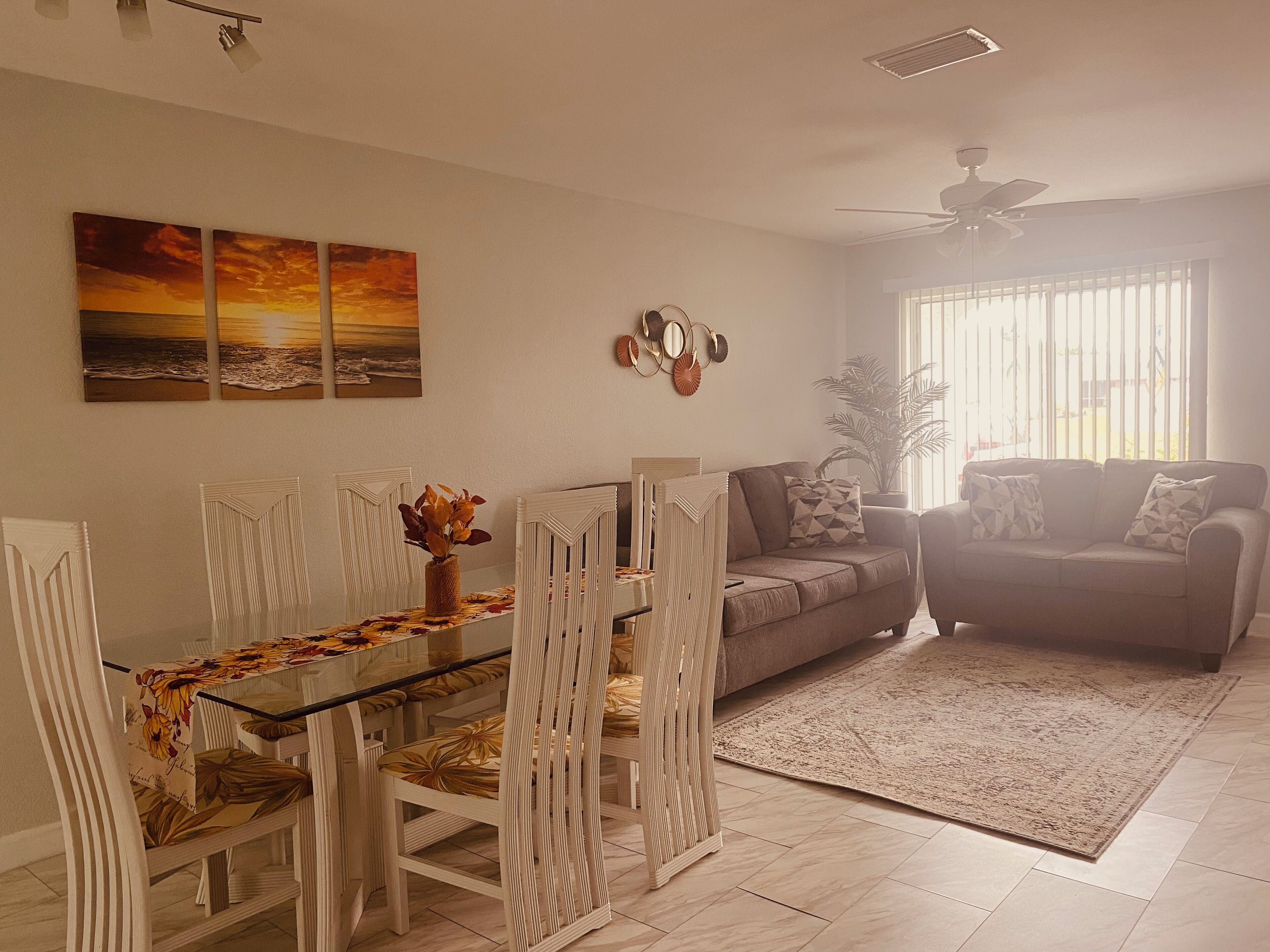 A dining area with a glass-top table, white chairs, and a vase with dried flowers. There is a sofa and a large window with blinds in the background.