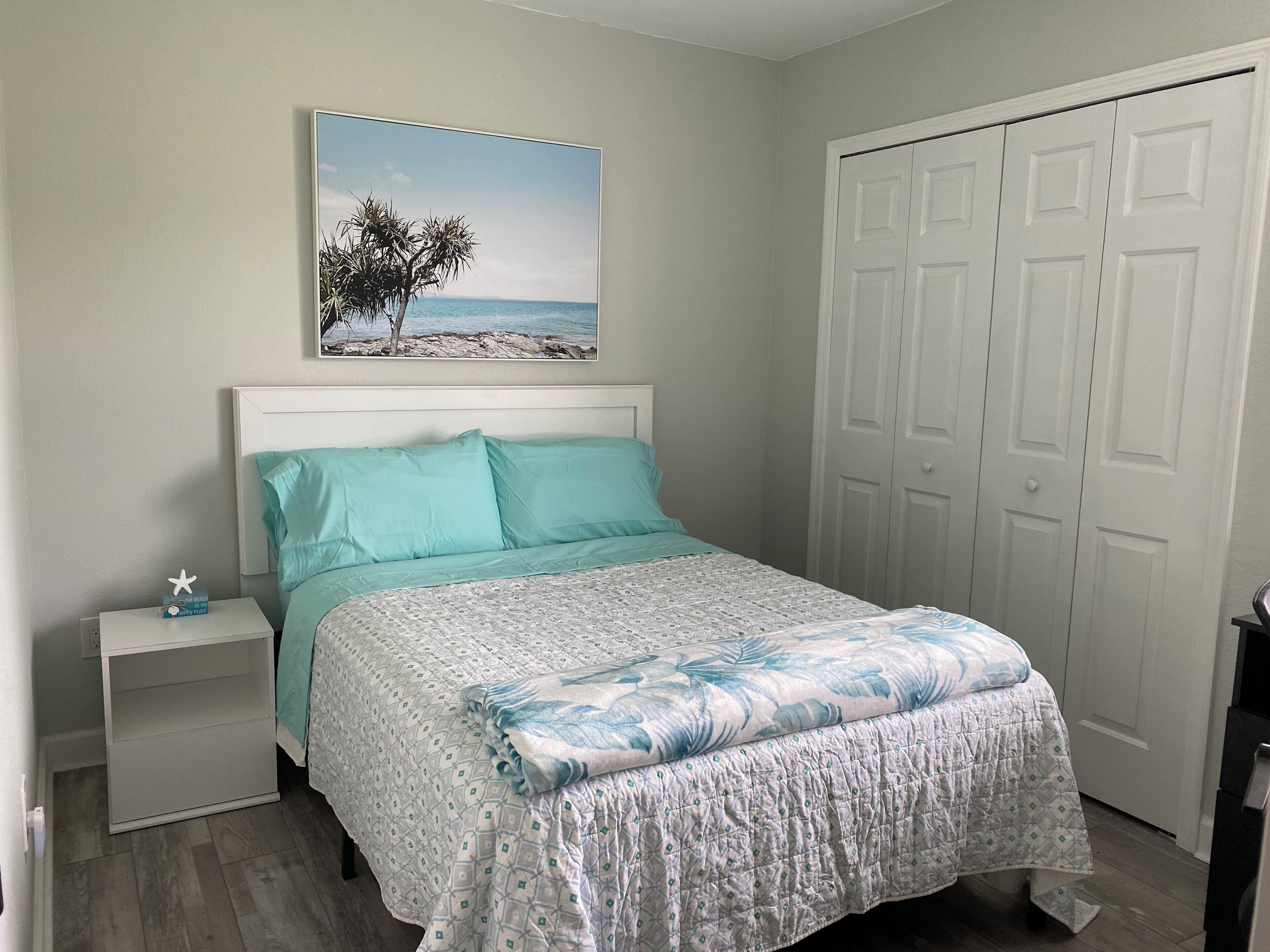 Une chambre à coucher avec un lit, une table de chevet, une armoire et un tableau sur le mur représentant un paysage de plage.