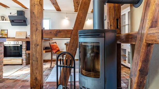 Un interior rústico con vigas de madera a la vista, un estufa de leña, un horno de ladrillo y un comedor con mesa y sillas de madera.