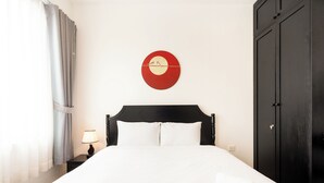 Una habitación de hotel con una cama, una mesita de noche con lámpara, un reloj de pared rojo y un armario oscuro.