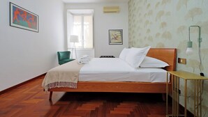 Une chambre d’hôtel bien rangée, avec un lit en bois, une chaise verte et une petite table sur laquelle se trouve une lampe.