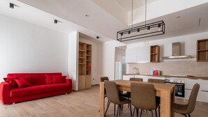 A modern living room with a red sofa, wooden dining table, and chairs. A kitchen with white cabinets and a stainless steel range hood is visible in the background. - Russu Suli Apartment 1 by Wonderful Italy (Siracusa)
