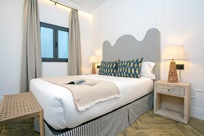 A neatly made bed with a striped headboard, two bedside tables with lamps, a small bench, and a window with curtains. - Genteel Home Lagar II (Seville)