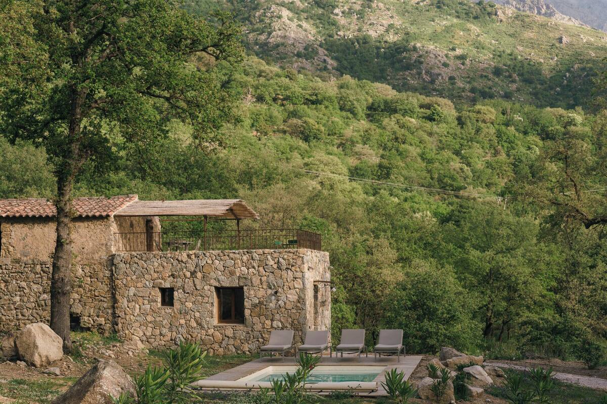 A stone building with a pool and lounge chairs, surrounded by lush greenery and hills.