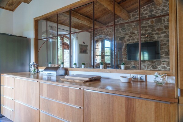 A kitchen with wooden cabinets, a large window, and a stone wall. - Domaine Acciola (Nessa)