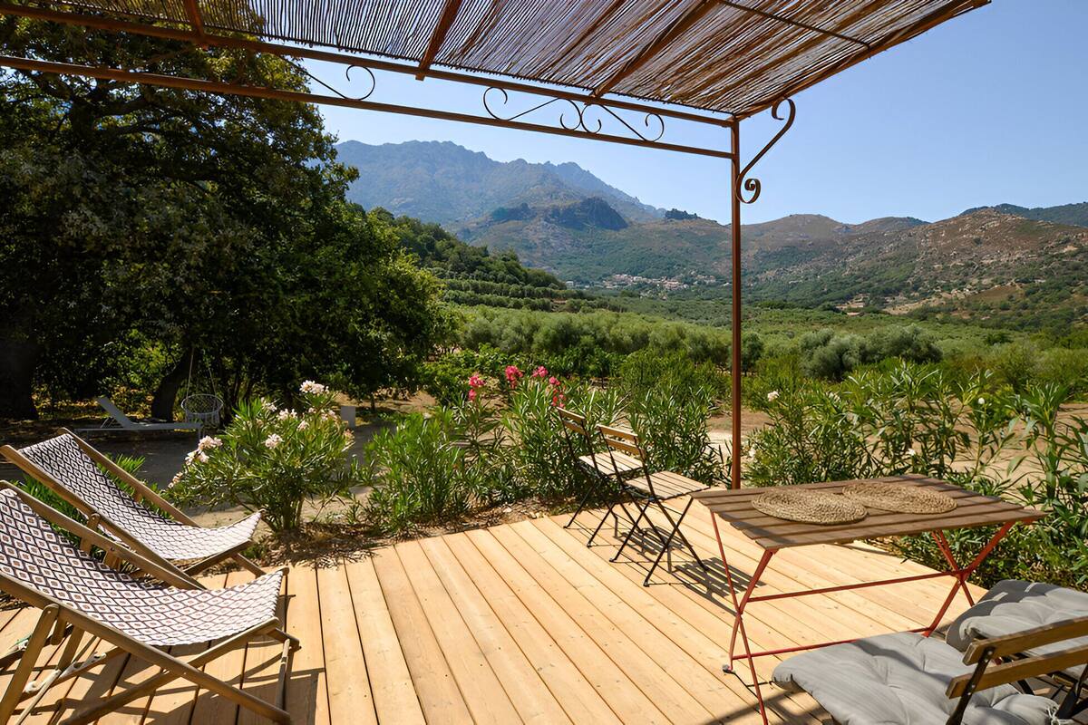 A wooden deck with lounge chairs and a table, overlooking a mountainous landscape with greenery.