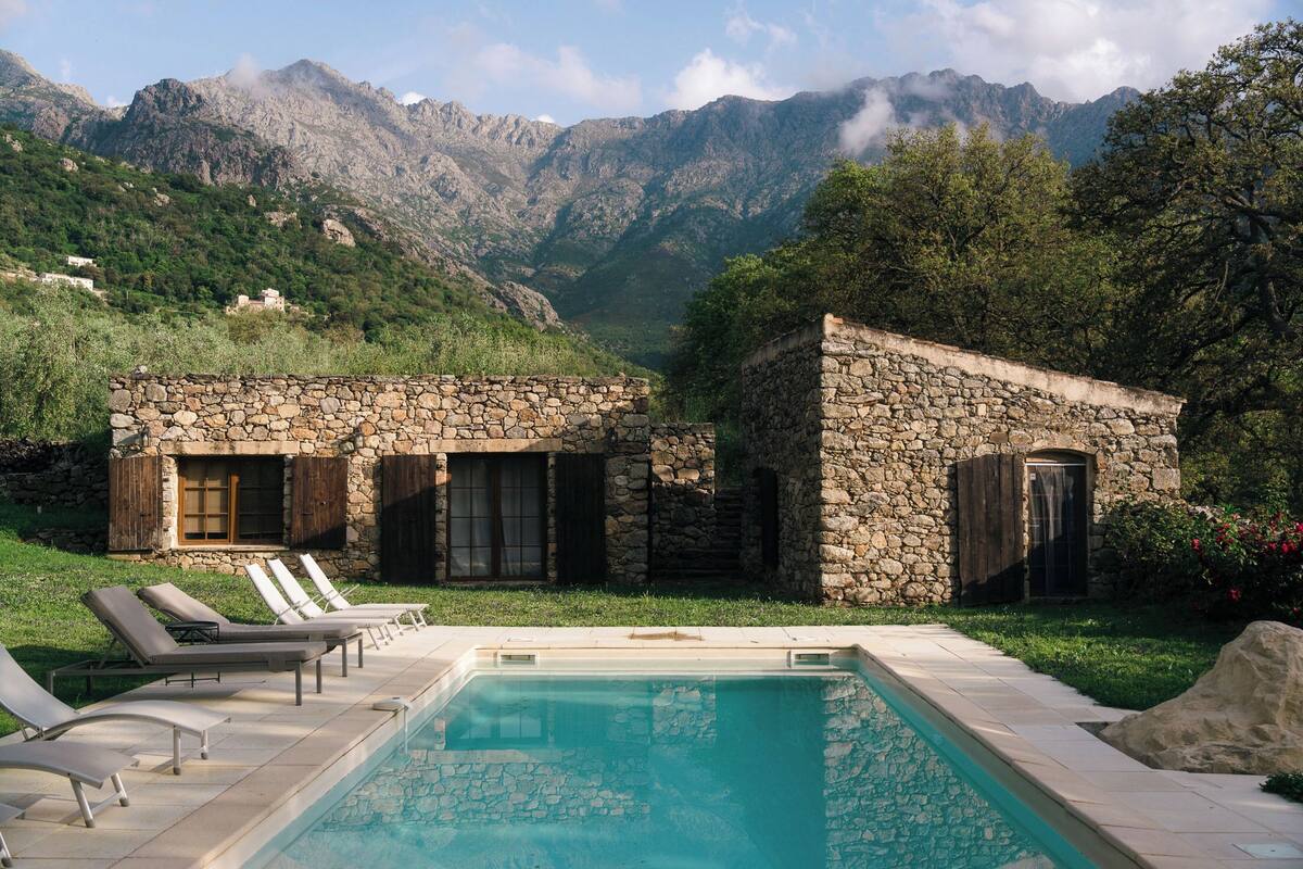 A stone villa with a pool, surrounded by mountains and greenery.