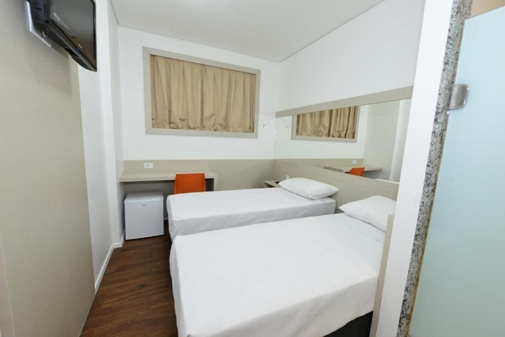 Een kleine, schone kamer met twee eenpersoonsbedden, een bureau, een stoel en een aan de muur bevestigde televisie.