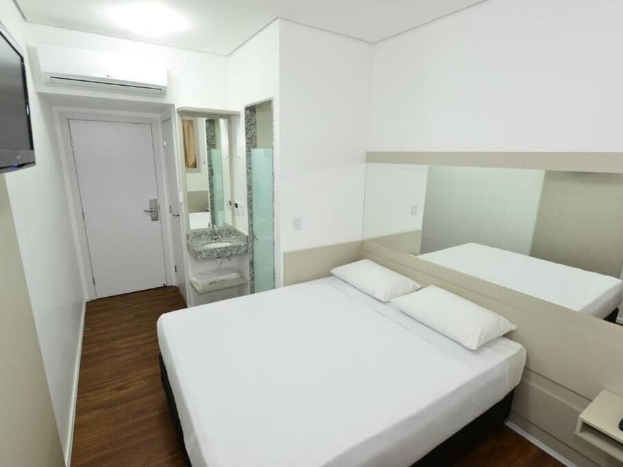 Quarto de hotel com cama, televisão, espelho e pequena mesa de cabeceira.