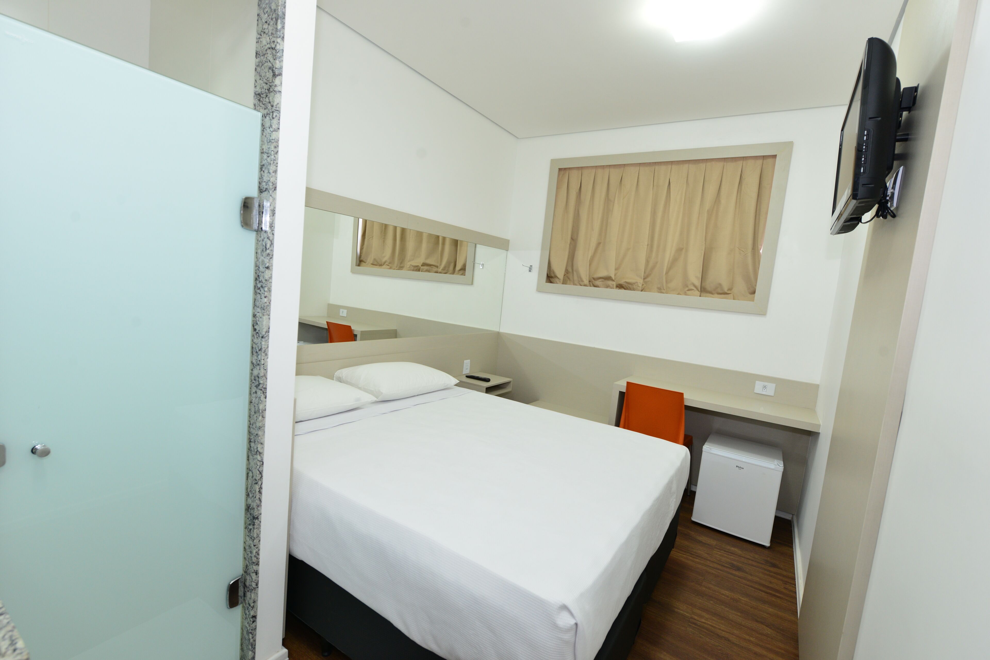 Une chambre d’hôtel équipée d’un lit, d’un bureau, d’une chaise, d’un petit réfrigérateur et d’un téléviseur fixé au mur.