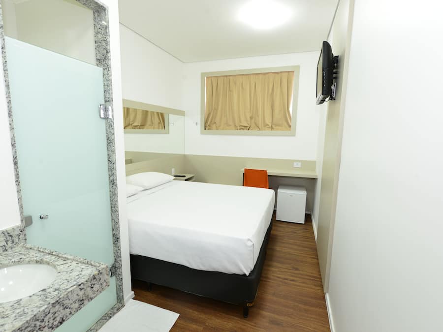 A hotel room with a bed, a small desk, a mirror, a window with curtains, and a sink.