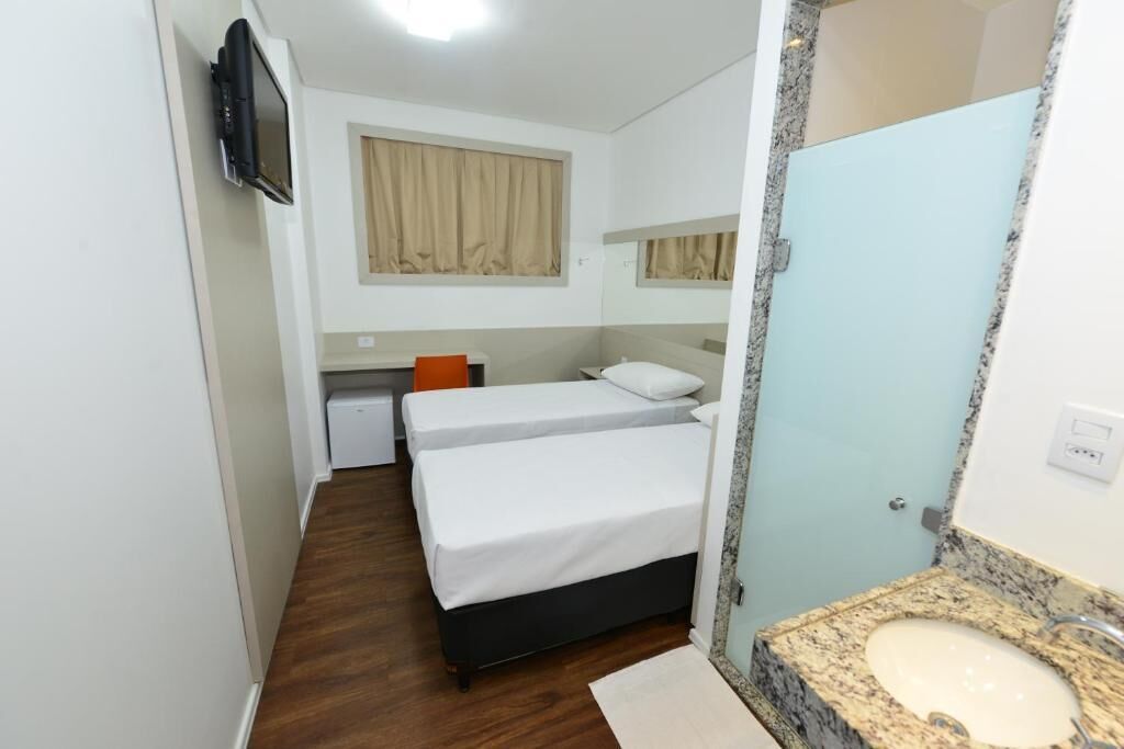 Habitación de hotel con dos camas individuales, un escritorio pequeño, televisor, baño con lavabo y espejo, y ventana con cortinas.