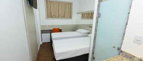 Habitación de hotel con dos camas individuales, un escritorio pequeño, televisor, baño con lavabo y espejo, y ventana con cortinas.