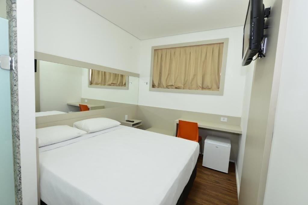 Una camera d'albergo con un letto, una scrivania, una sedia, un piccolo frigorifero e una TV fissata al muro.
