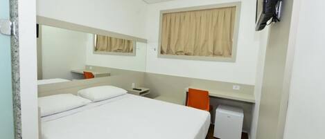 Una camera d'albergo con un letto, una scrivania, una sedia, un piccolo frigorifero e una TV fissata al muro.