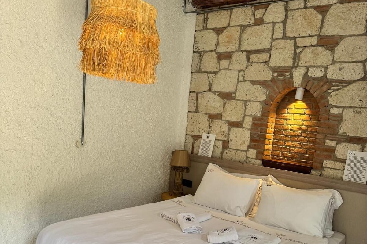 A bed with white linens, a hanging rattan light fixture, a stone wall with a brick arch, and a wooden headboard.