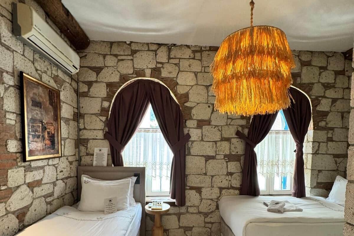 A room with two beds, a stone wall, a hanging light fixture, a small table, and a window with curtains.