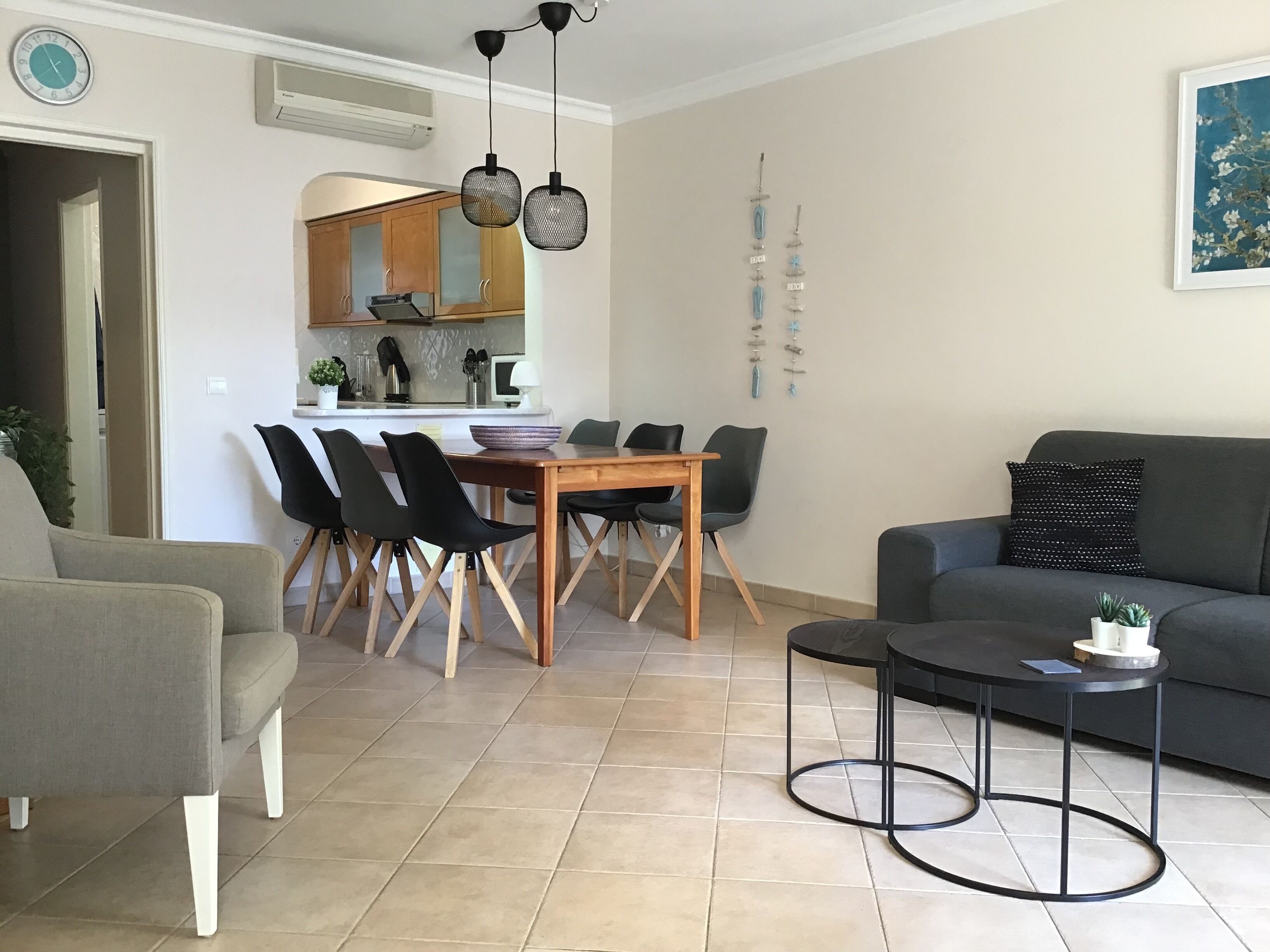 A modern living room with a grey sofa, a wooden dining table with chairs, a small black side table, and a grey armchair.