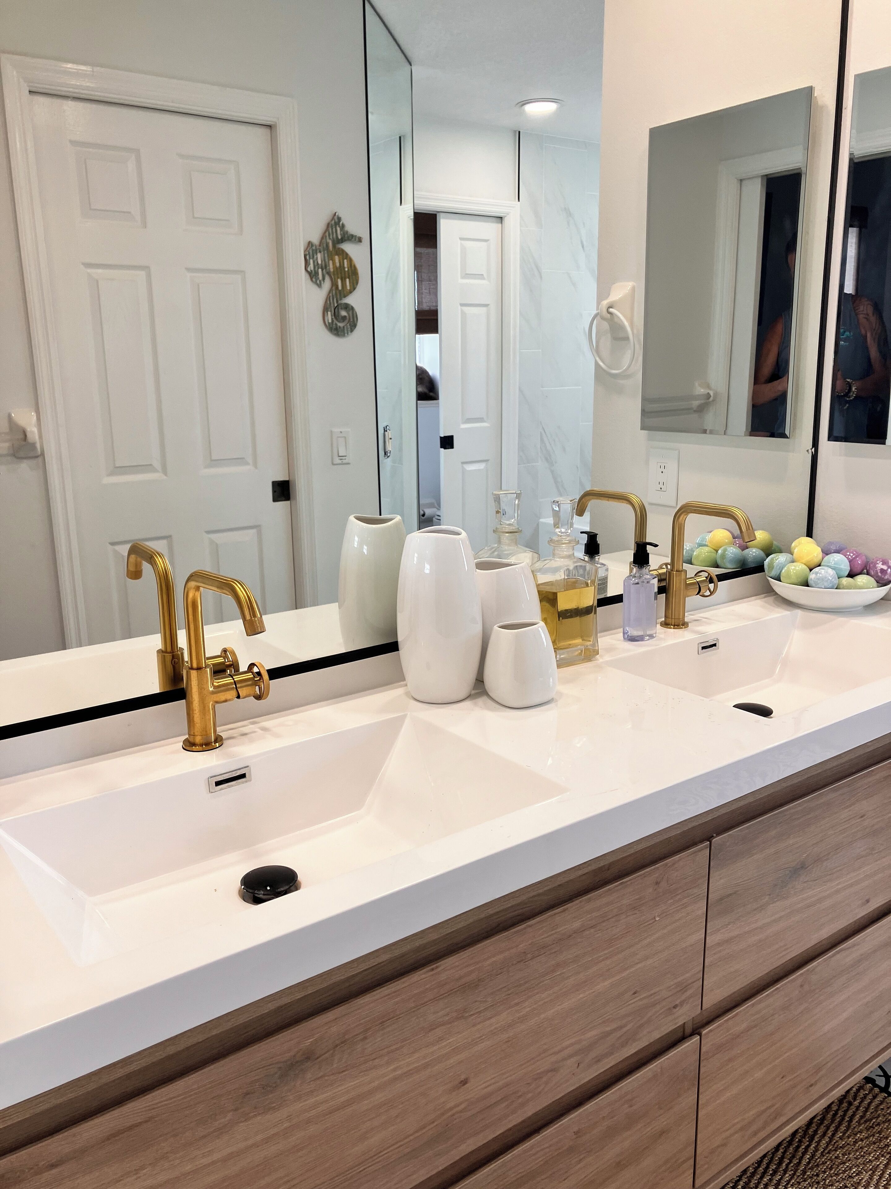 A modern bathroom with a double sink, gold faucets, and a large mirror. There are toiletries on the counter and a towel rack on the wall.
