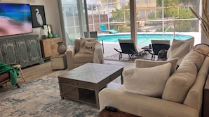 A living room with a large TV, comfortable seating, and a view of a pool. - The Windsor @ Englewood Beach (Englewood)