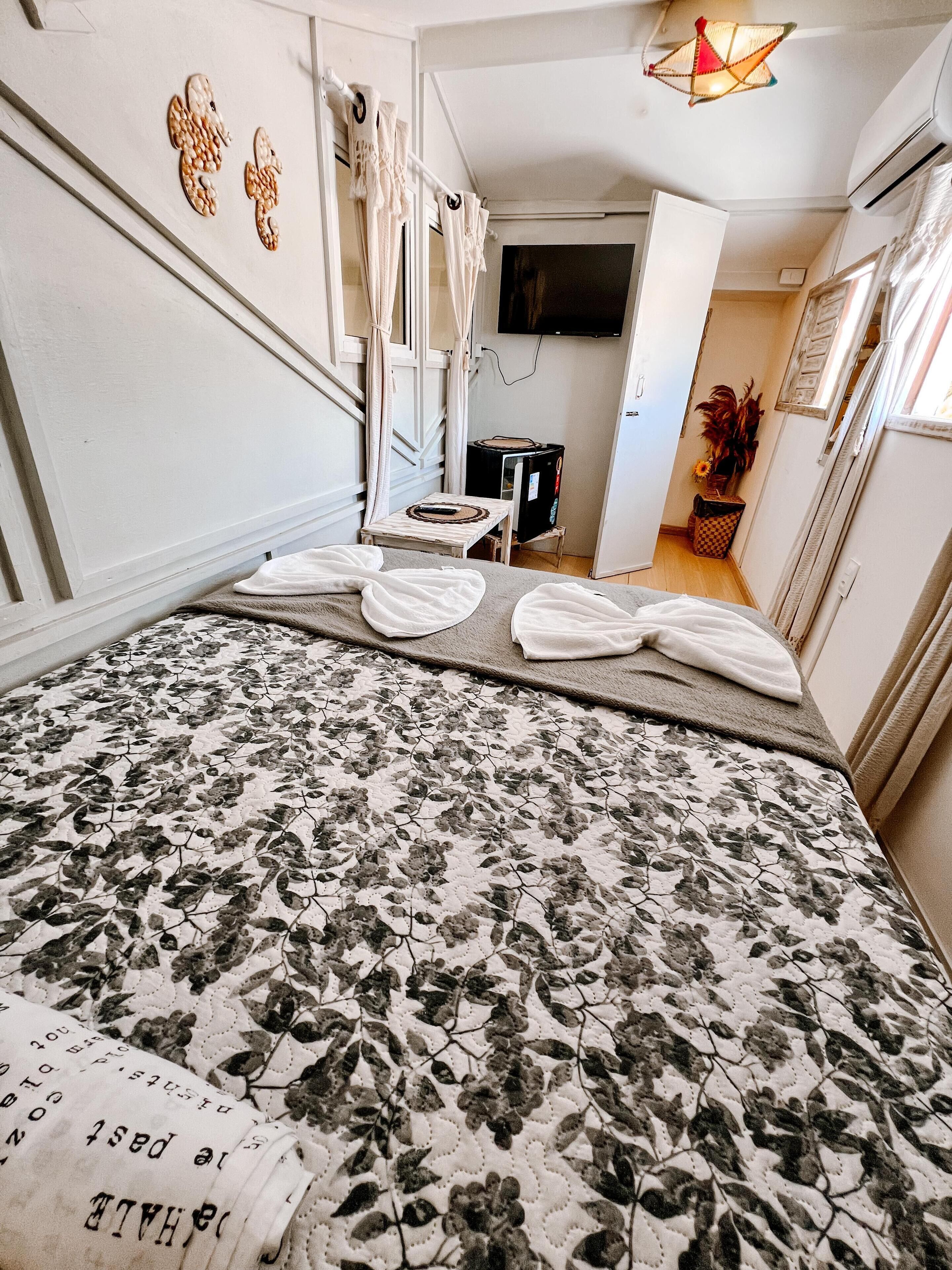 Habitación de hotel con una colcha de estampado floral, una mesita con un control remoto, un televisor montado en la pared y una planta en maceta en la esquina.