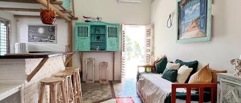 A room with a kitchen area, a bed with pillows, a hanging light fixture, and a painting on the wall.