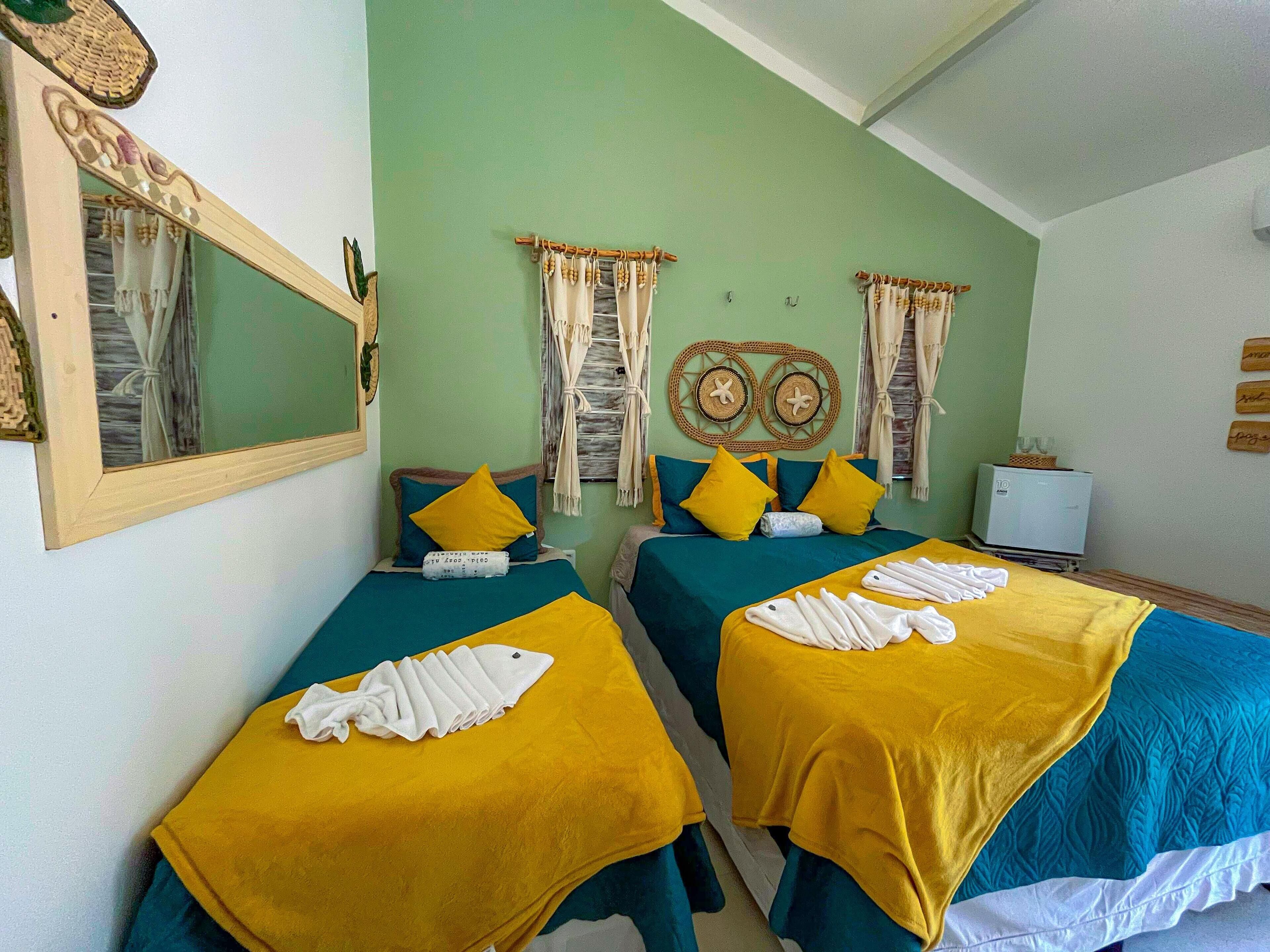 Habitación con dos camas, cada una con ropa de cama amarilla y azul, un espejo, un refrigerador pequeño y una pared decorada.