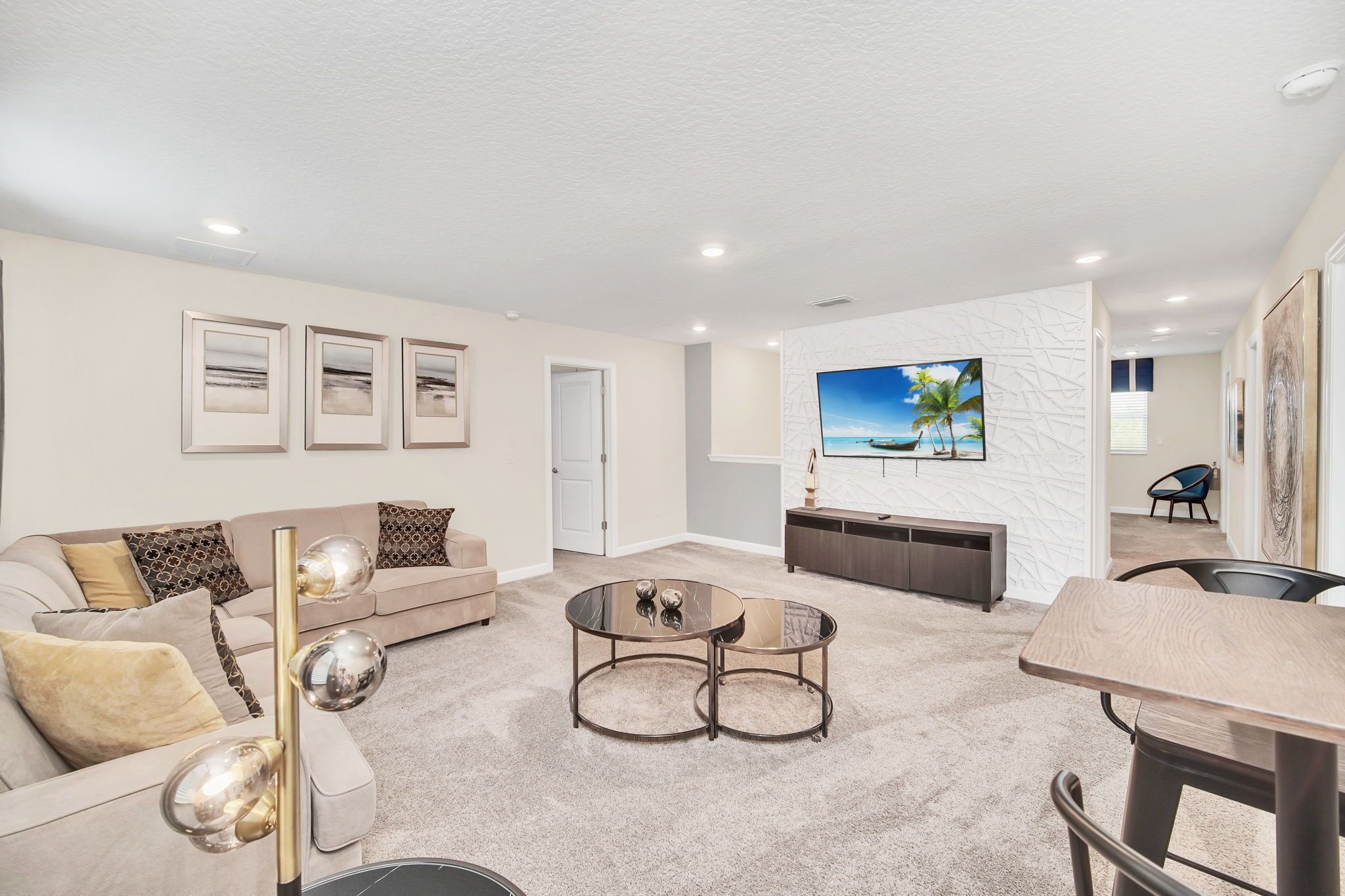 A modern living room with a beige sofa, a glass coffee table, and a flat-screen TV mounted on a white textured wall.