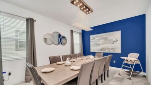 Une salle à manger avec une grande table en bois, dressée pour six personnes, entourée de six chaises, d’une chaise haute pour bébé et d’un lustre.