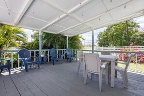 Uma área de estar coberta ao ar livre com cadeiras e uma mesa, cercada por vegetação e um muro branco.