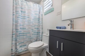 Casa de banho com sanita branca, armário cinzento, toalha azul e cortina de chuveiro estampada.